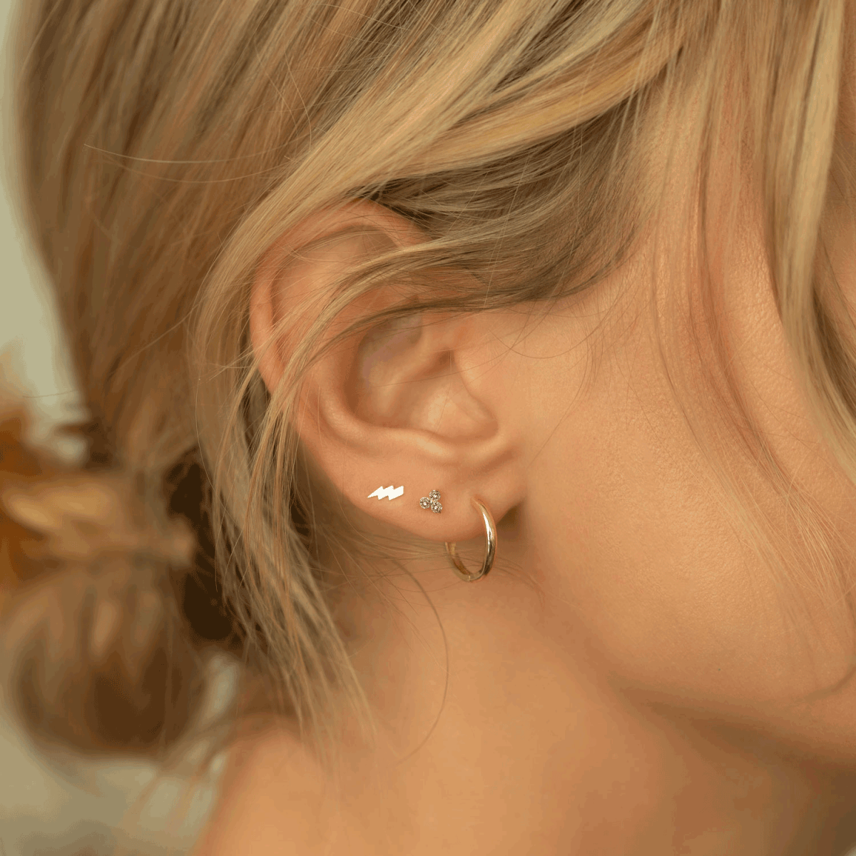 Close up of woman's ear featuring 14K Gold Gemstone Clover Studs Amethyst CZ Crystal paired with lightning bolt stud and gold hoop earring
