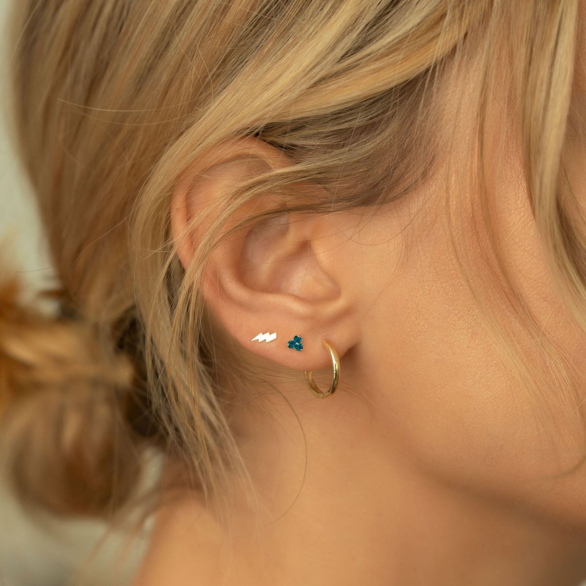 Close up of woman's ear featuring 14K Gold Gemstone Clover Studs Turquoise CZ Crystal, lightning bolt stud, and gold hoop earring