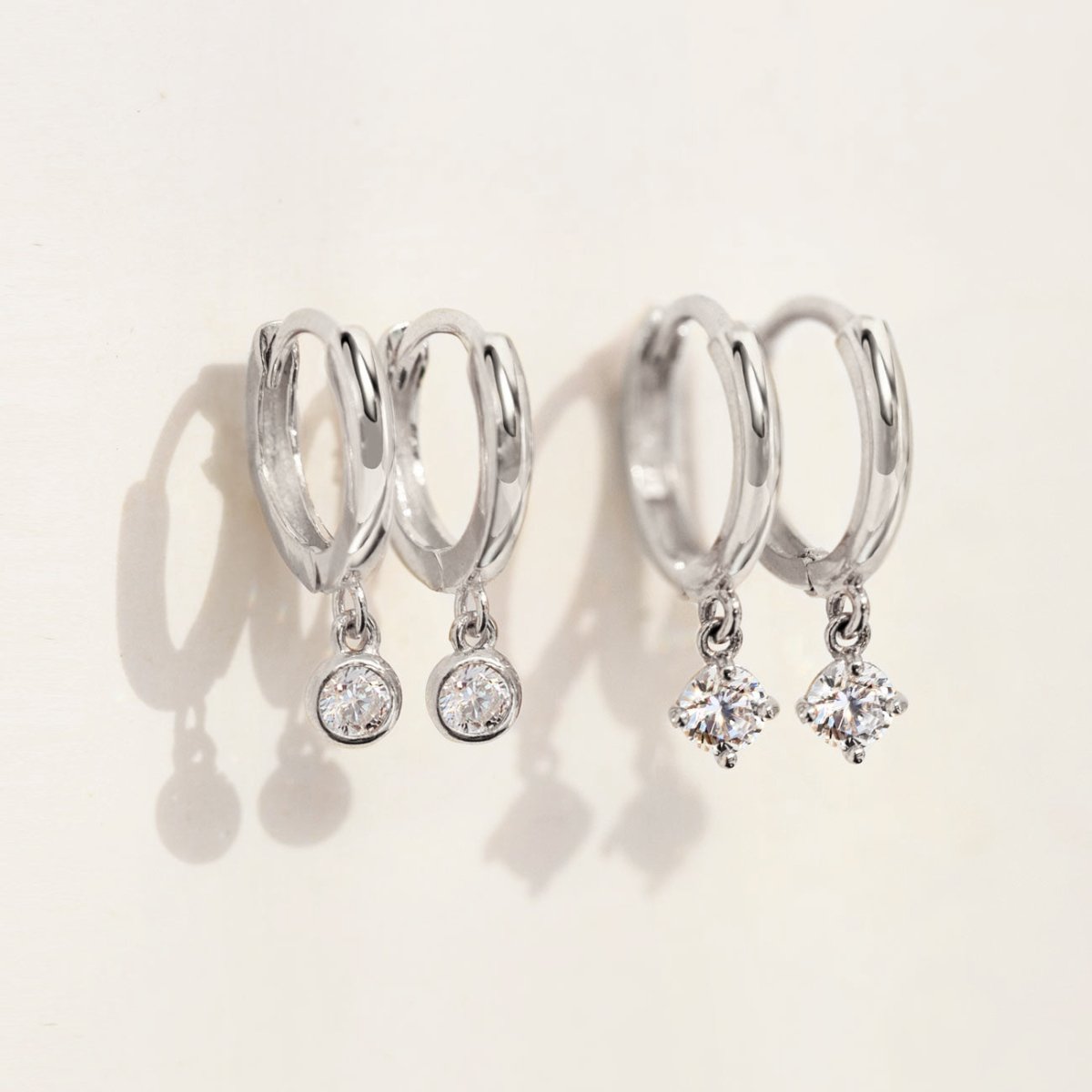 Sterling silver bezel and prong-set white zirconia dangle huggie hoops, shown on a cream background