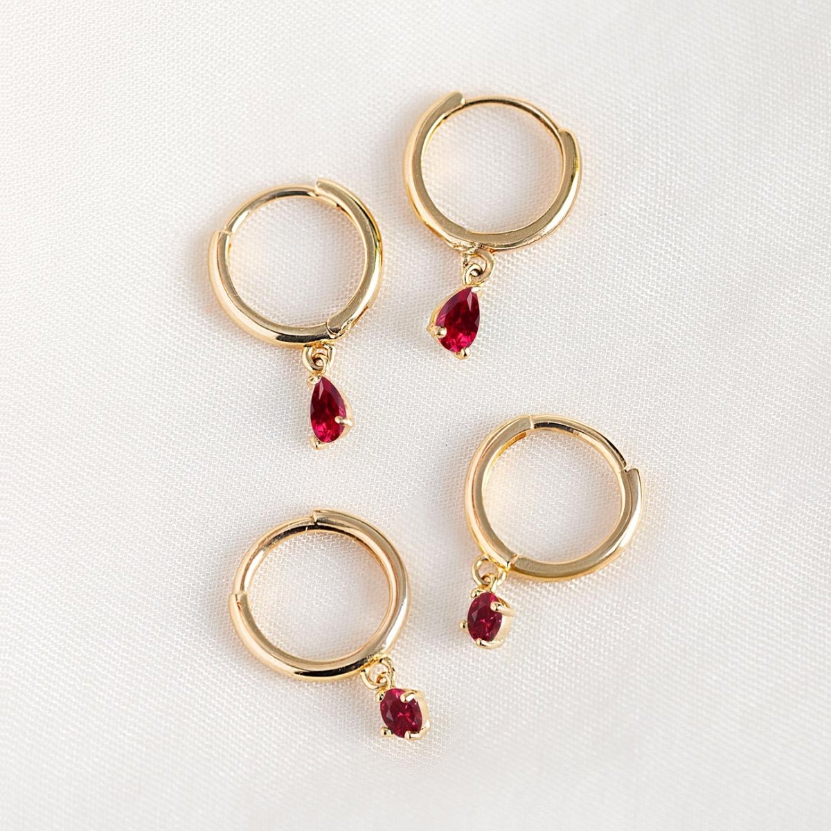 14K gold huggie hoops with pear-shaped red ruby drops, shown loose on a cream background