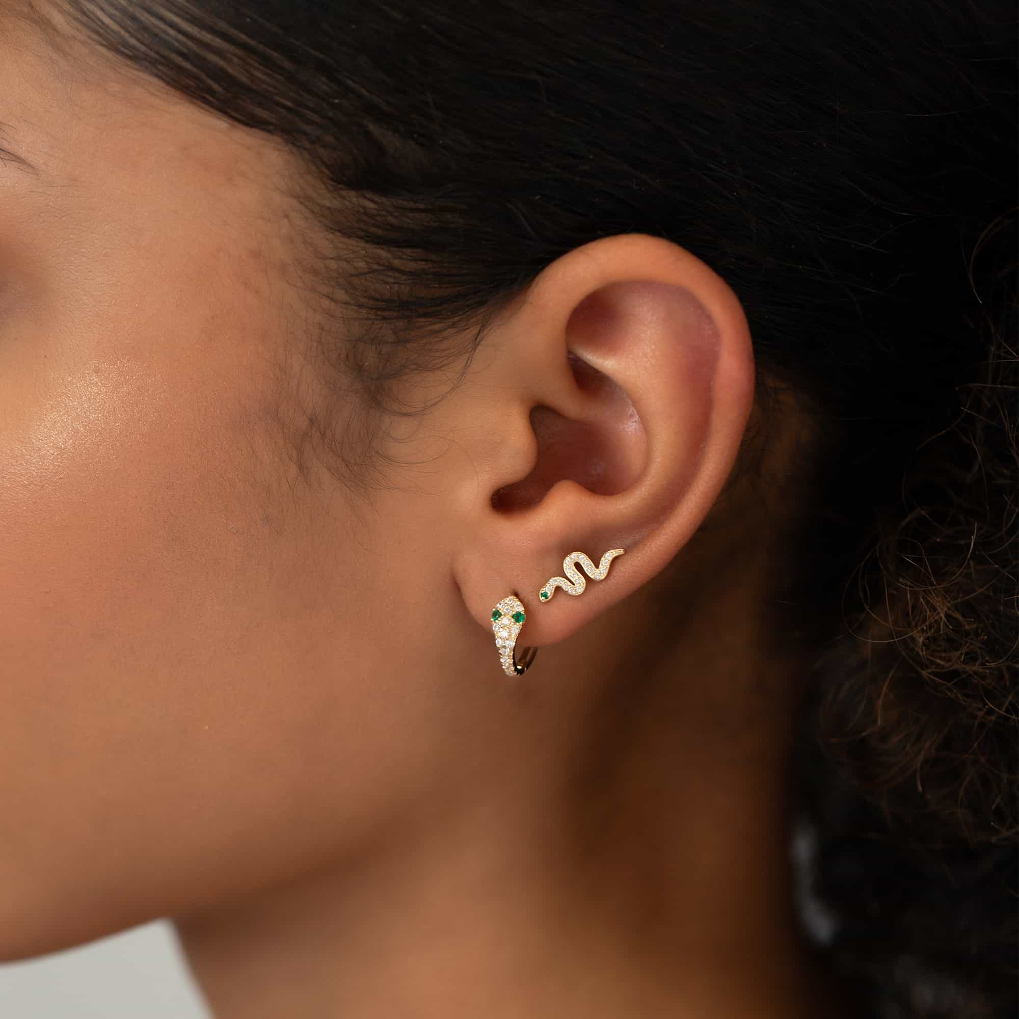14K gold pavé snake huggie hoop with green eyes, shown on a model's lobe with a snake stud above