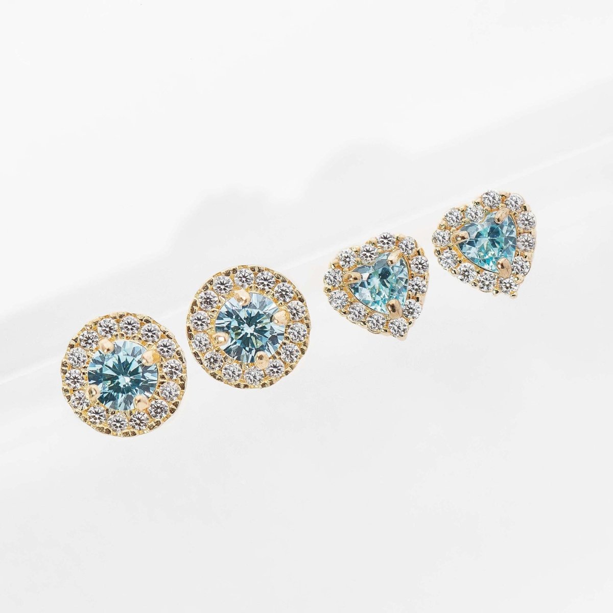 14K gold aquamarine halo studs in round and heart cuts, laid in a row on a glossy white surface