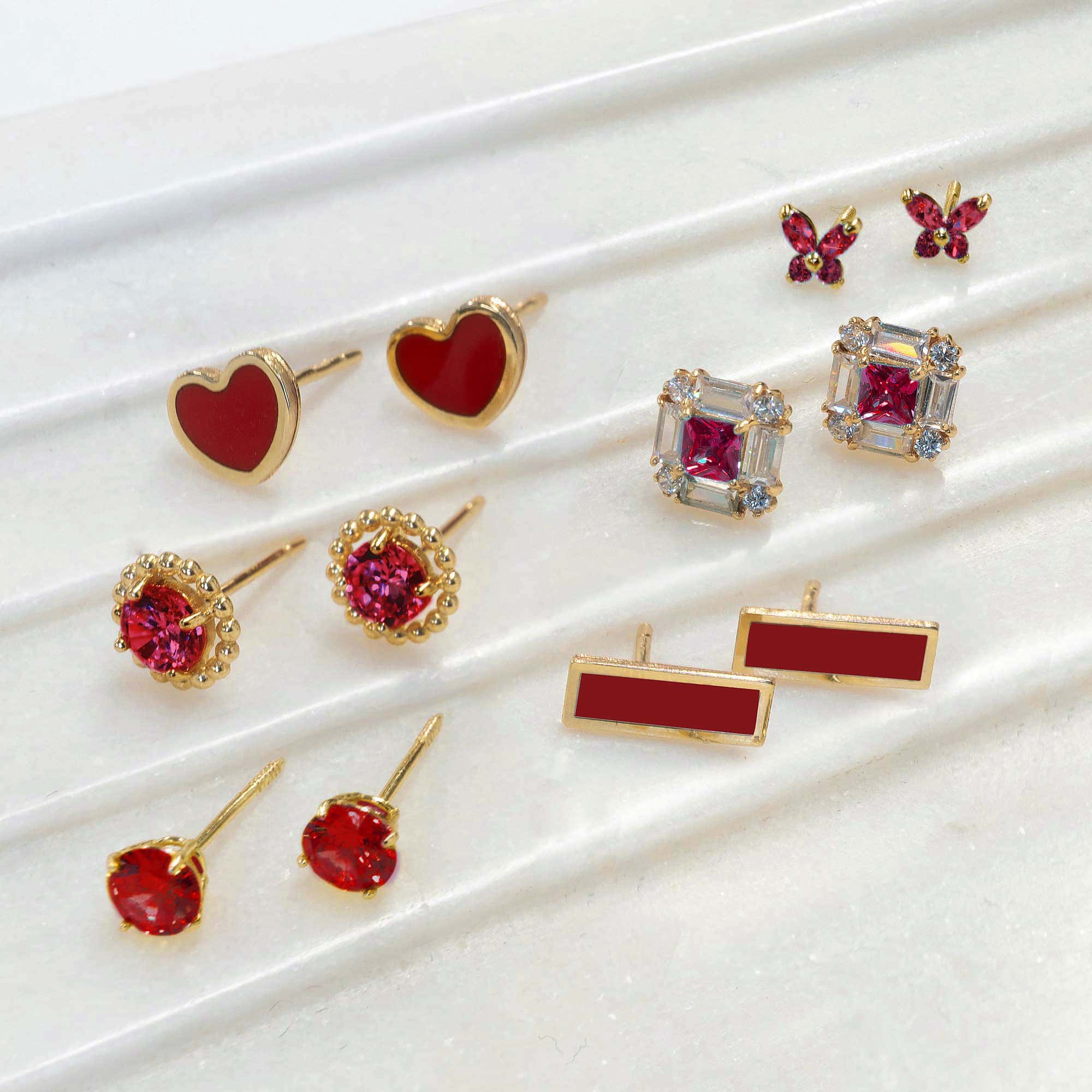 An assortment of gold red-stone and enamel stud earrings in heart, round, butterfly, square and bar shapes, on white