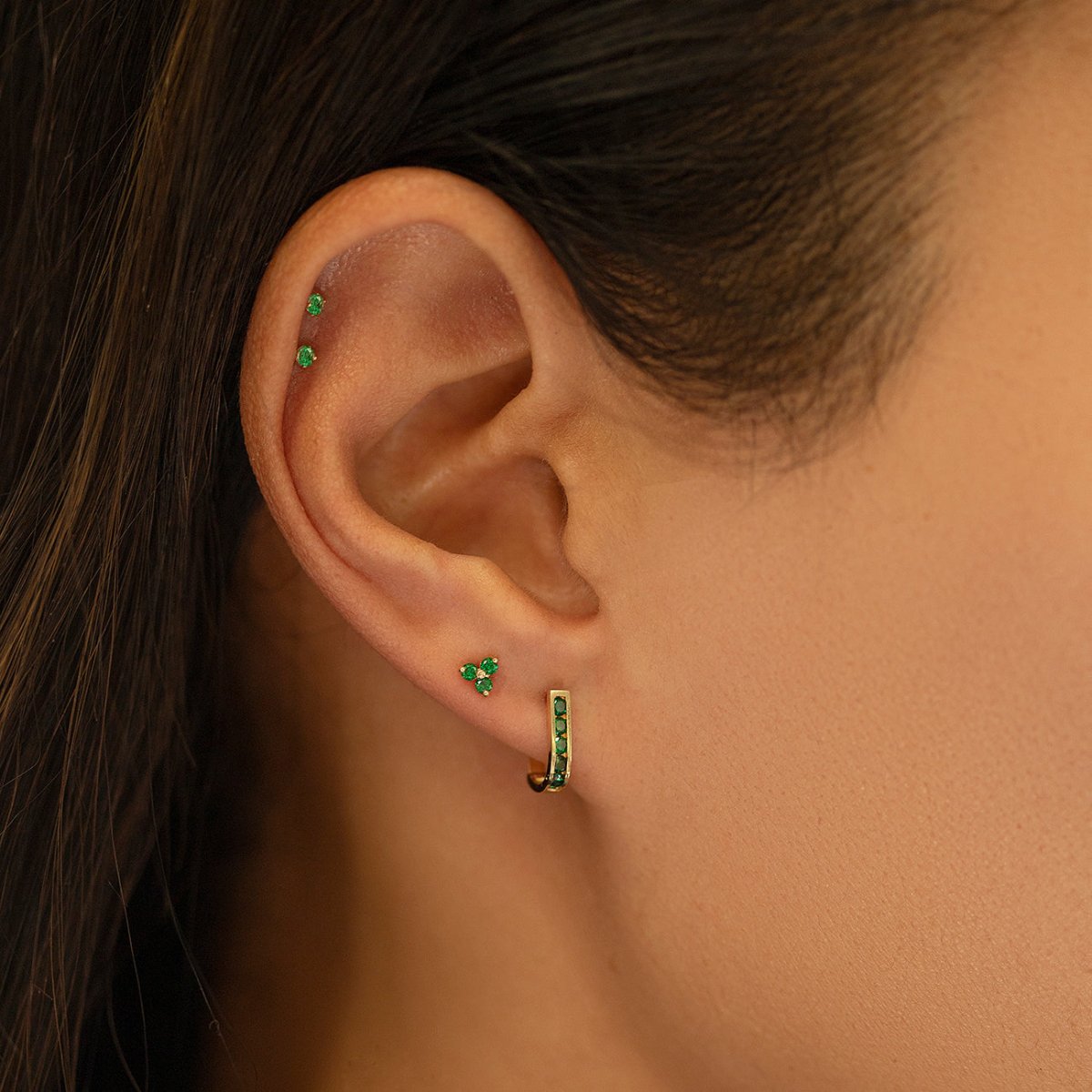 14K gold emerald huggie hoop with green stones, shown on a model's lobe in an ear stack