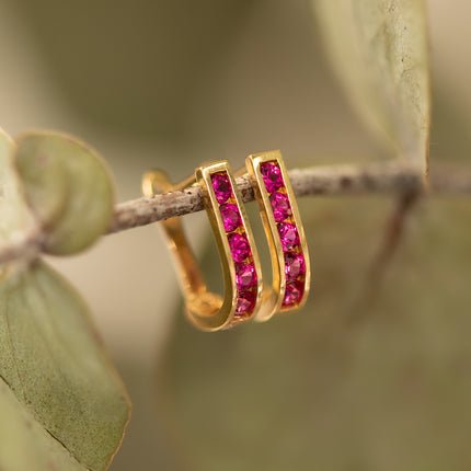 14K gold ruby huggie hoops with a row of red stones, resting on a branch
