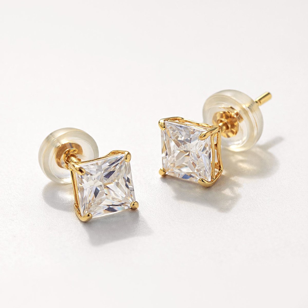 14K gold princess-cut studs with square clear stones and silicone backs, close-up detail on a cream background