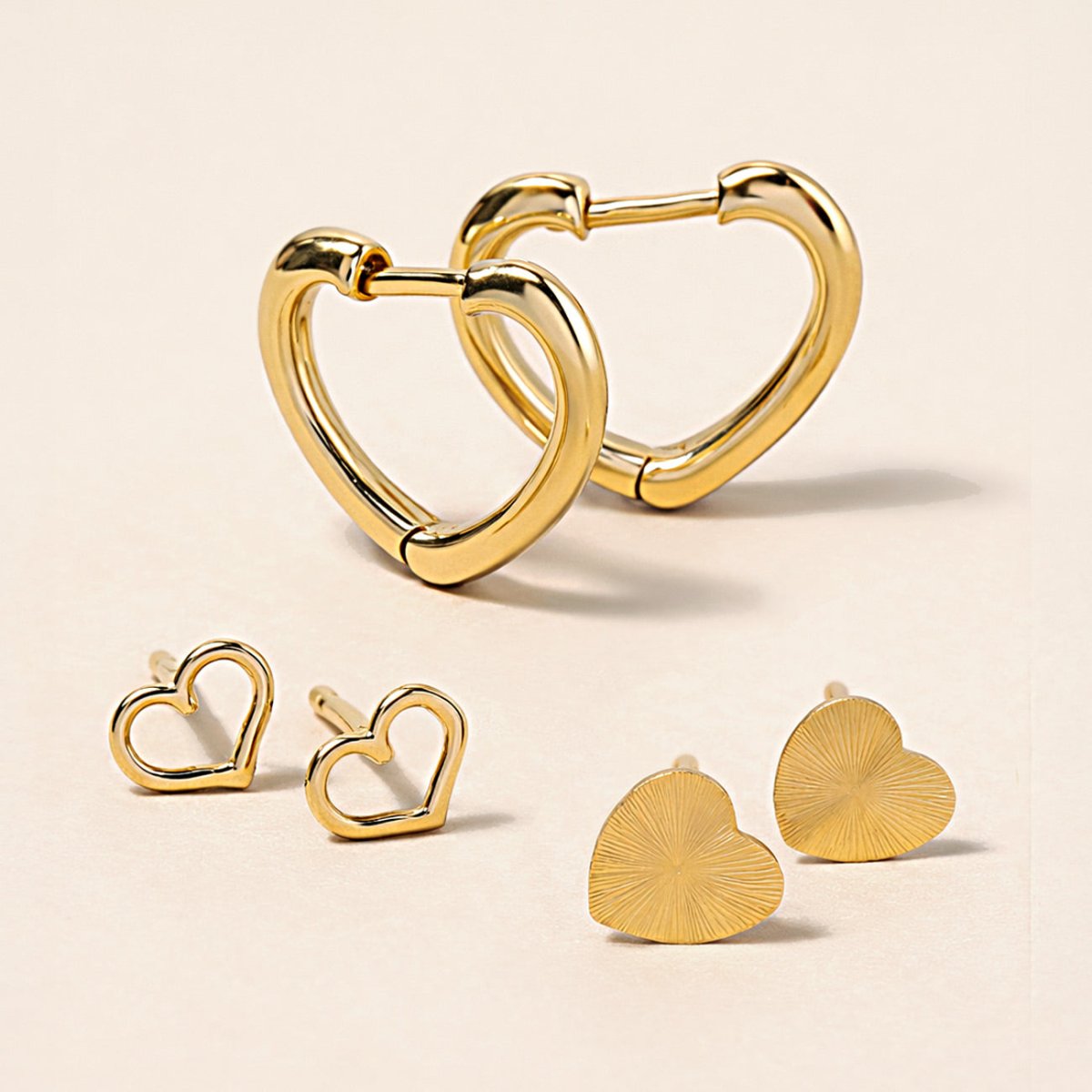 Gold heart earrings, including huggie hoops, open-heart studs and ridged heart studs, on a cream background