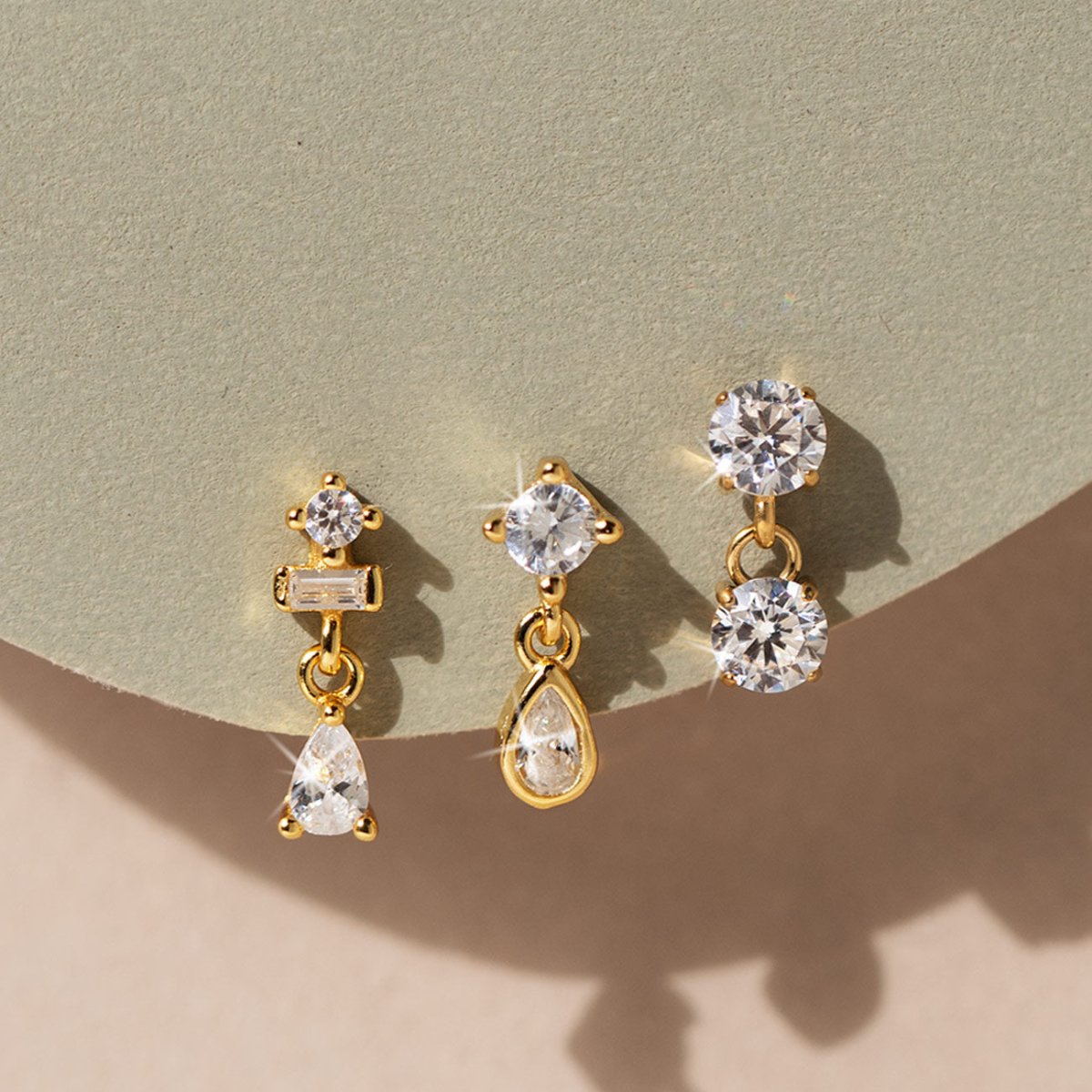Three gold dangle flat back studs with clear stones, on a sage and tan surface