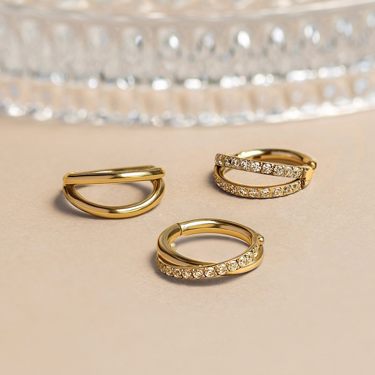 Three gold clicker hoops with double-bar and criss-cross pavé styles set with clear stones, on a cream surface
