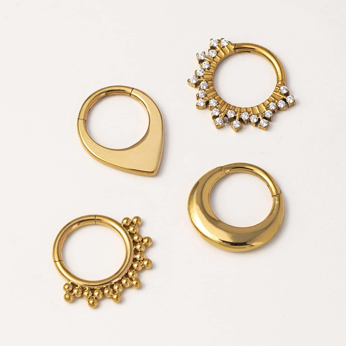 Four gold clicker hoops in teardrop, pavé clear-stone, beaded and crescent styles, on a cream background