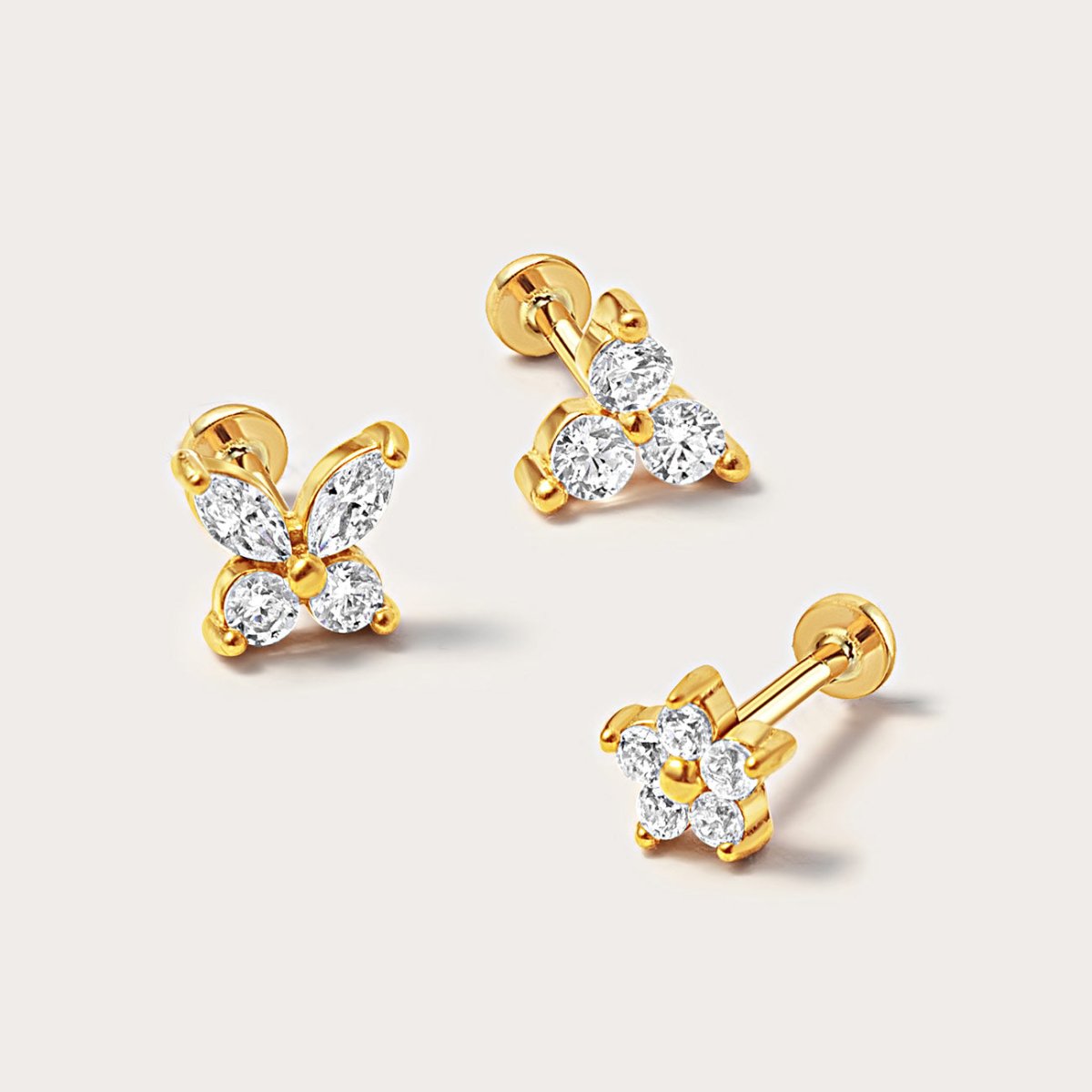 Three gold vermeil flat back studs with clear white zirconia in butterfly and flower shapes, on a cream background