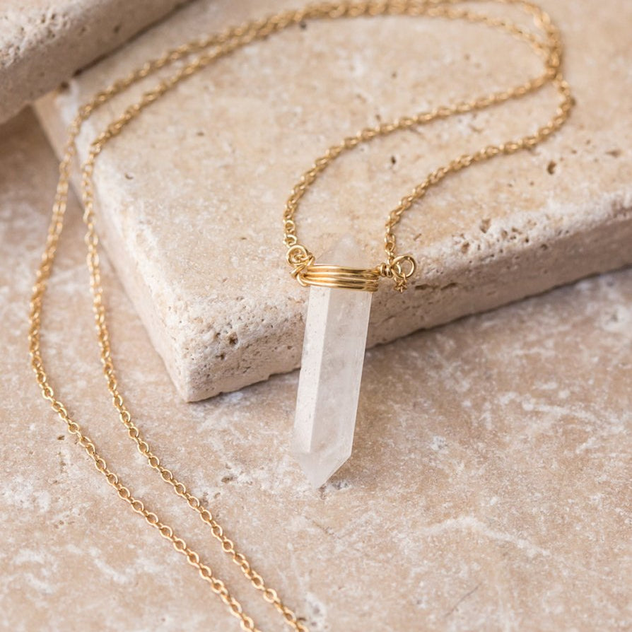 Gold vermeil necklace with a pointed clear quartz pendant, laid on white marble