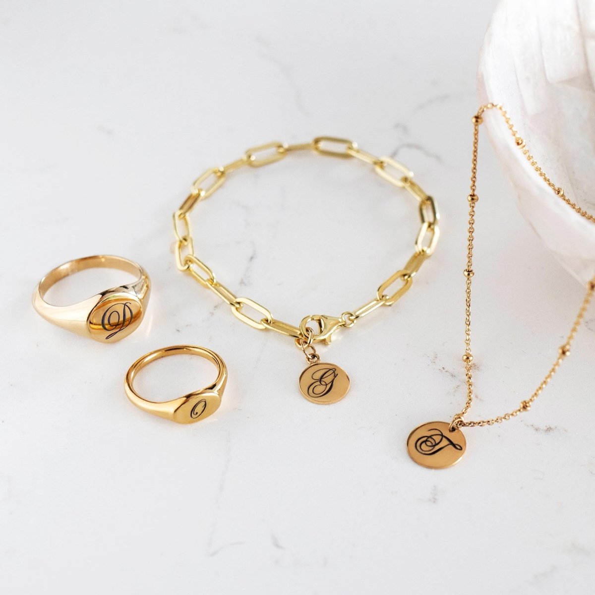 Gold signet rings, paperclip bracelet and disc necklace with engraved initials, on white marble