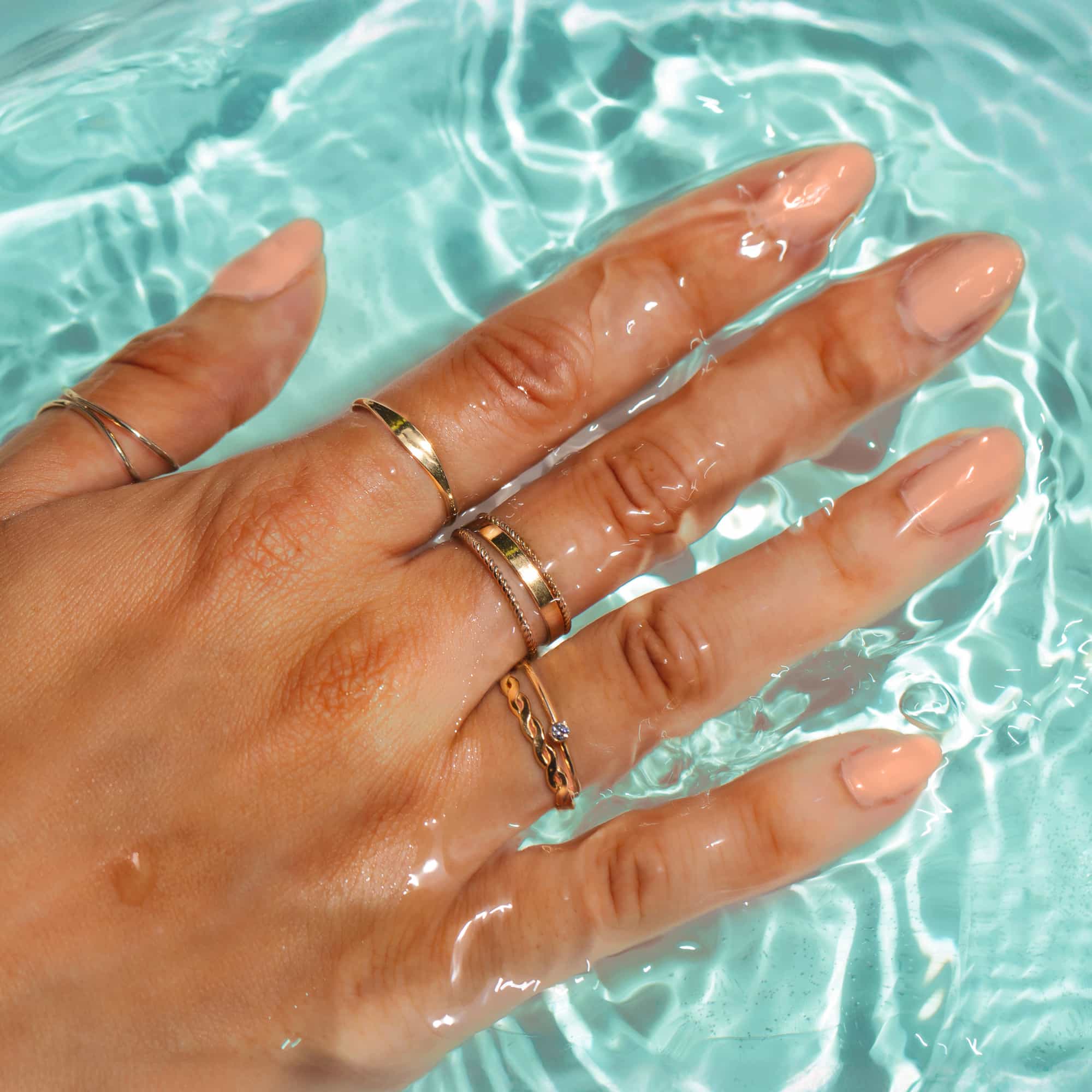 Stacked gold bands and a twisted rope ring on a hand resting in clear blue water