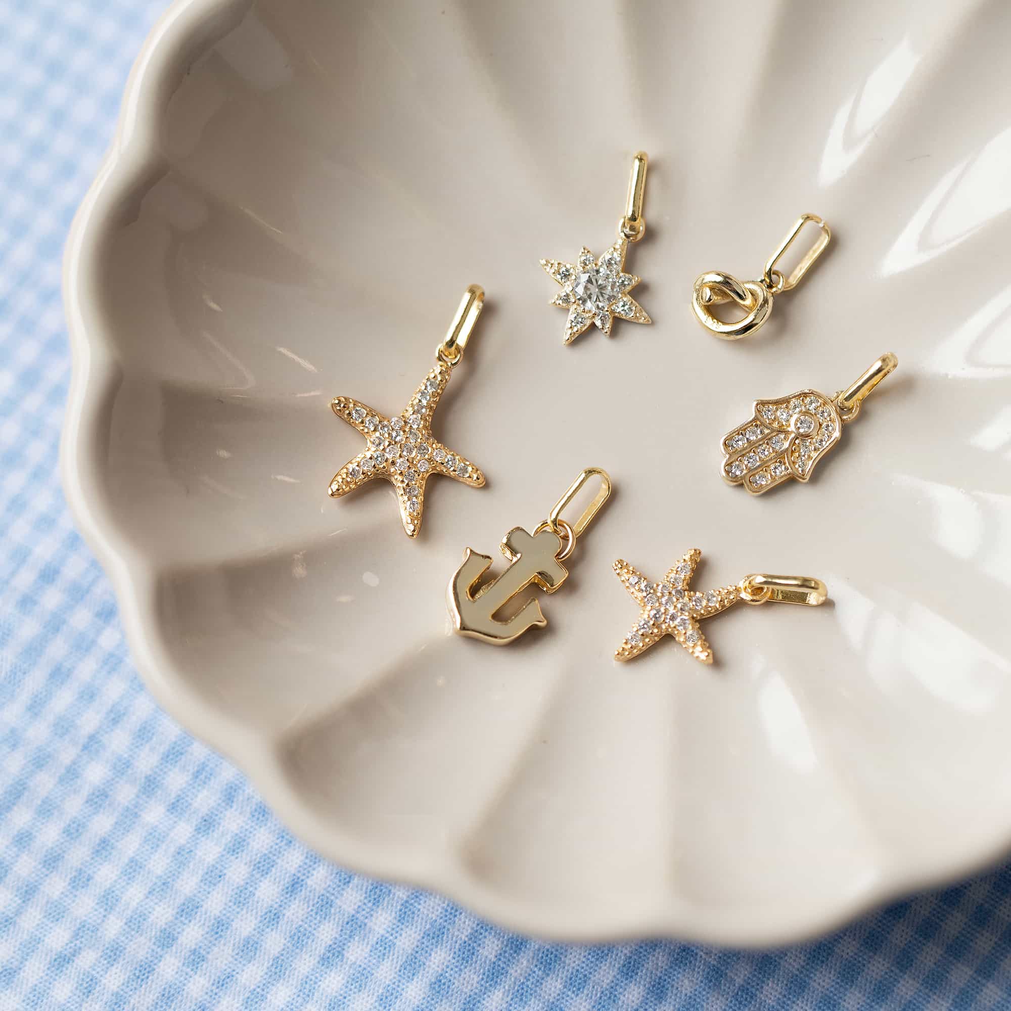 Assorted gold charms including starfish, star, hamsa and anchor in a shell dish on blue gingham