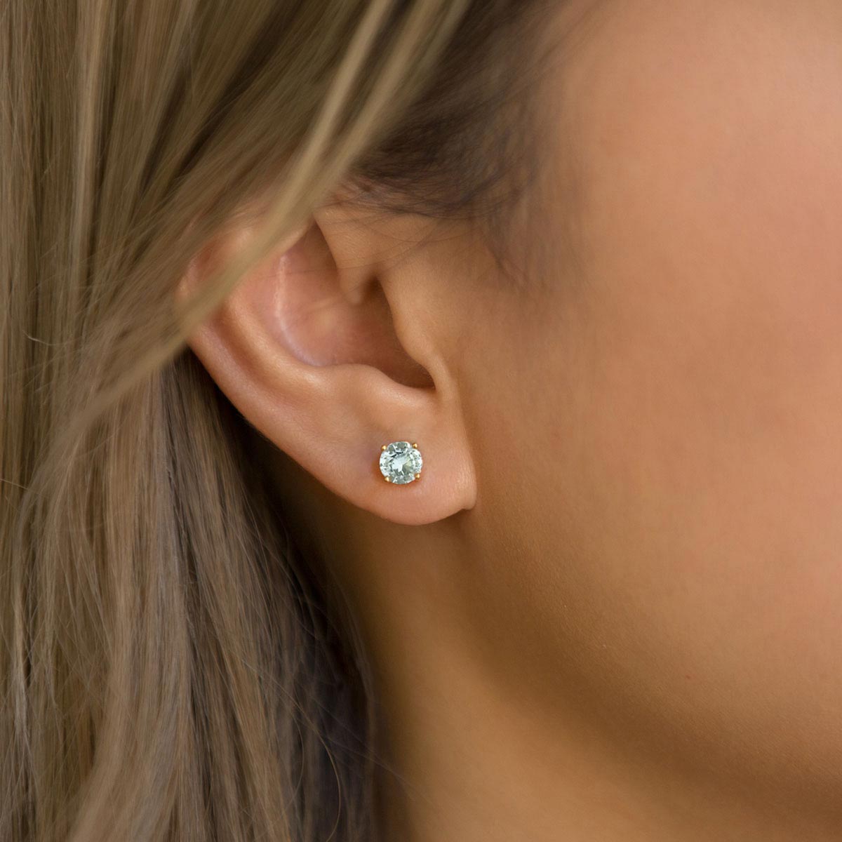 14K gold round aquamarine stud earring with a pale blue stone, shown on a model's ear