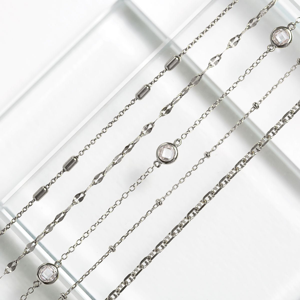 Sterling silver chain chokers with clear bezel-set stone stations, close-up detail over a glass surface