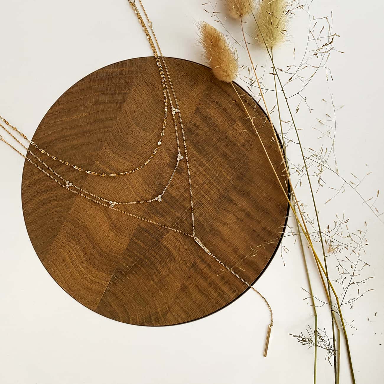 Layered gold necklaces with clover clusters of clear stones, laid on a wood disc with dried grasses