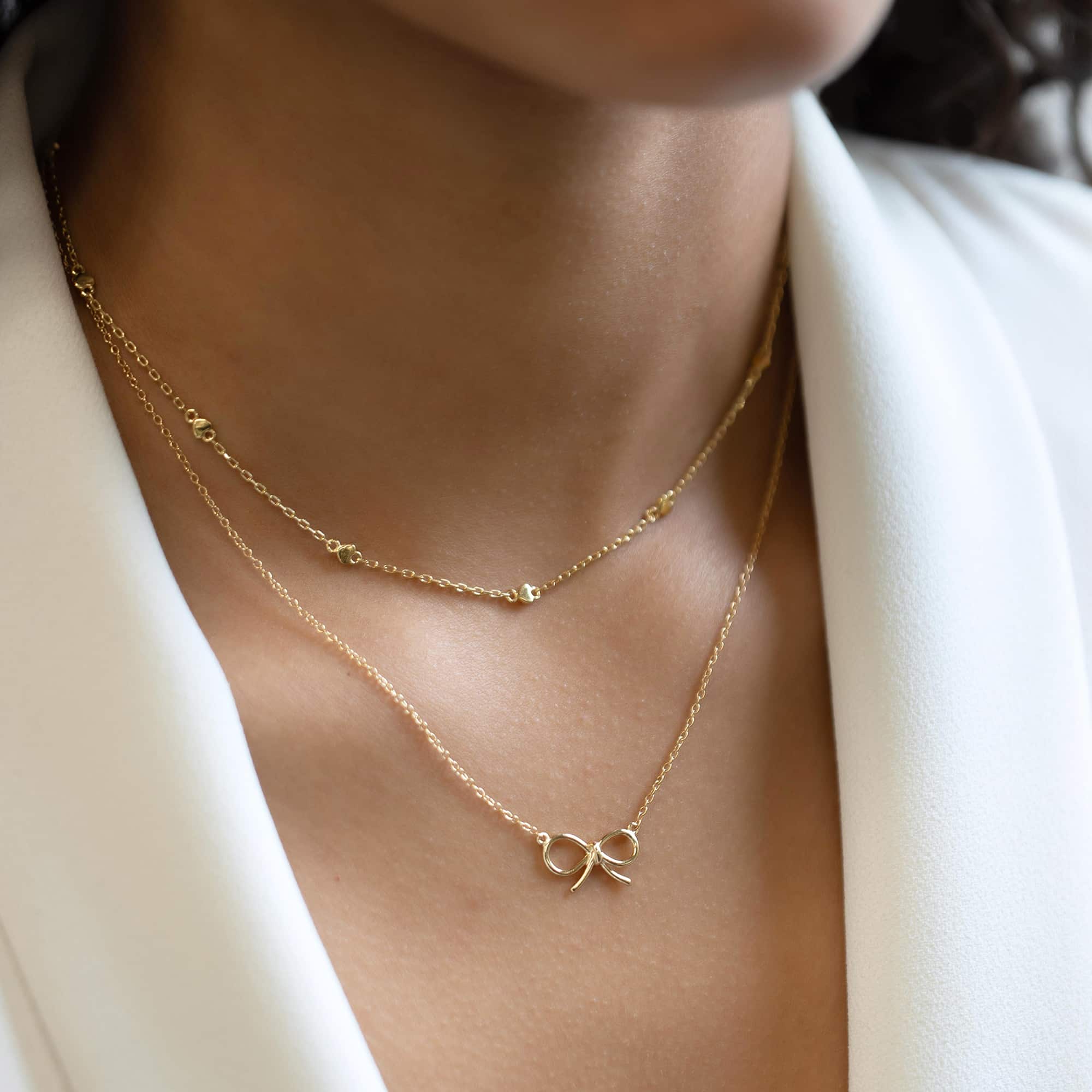 Layered gold necklaces, including a beaded chain and a bow pendant, on a model's neckline