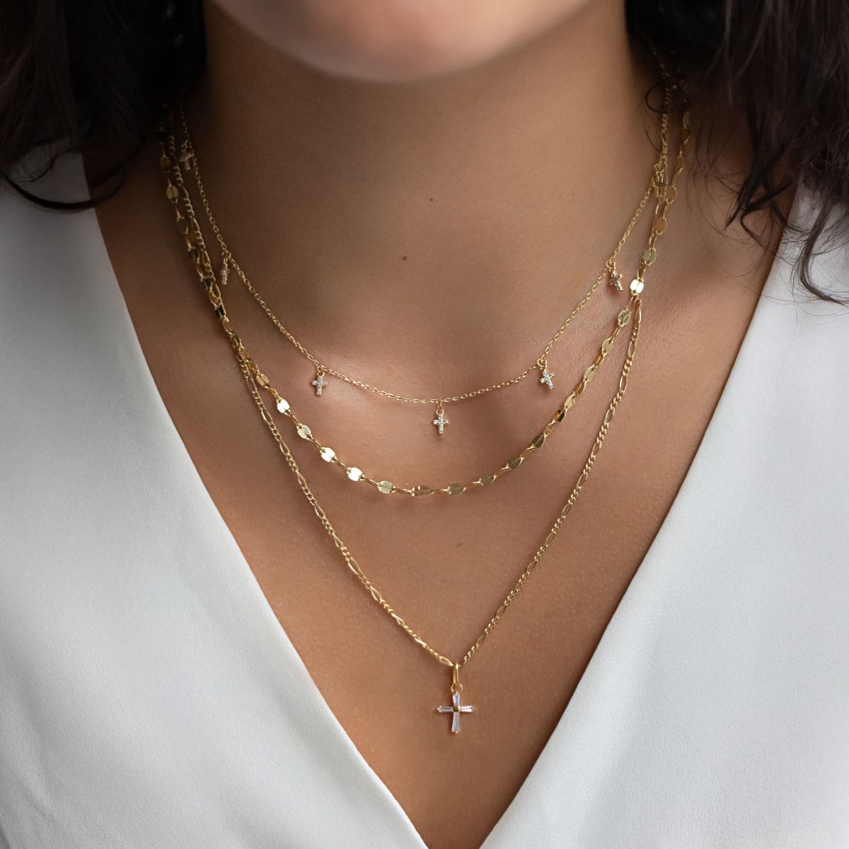 Gold cross charm pendant with clear baguette stones, shown layered on a model's neckline