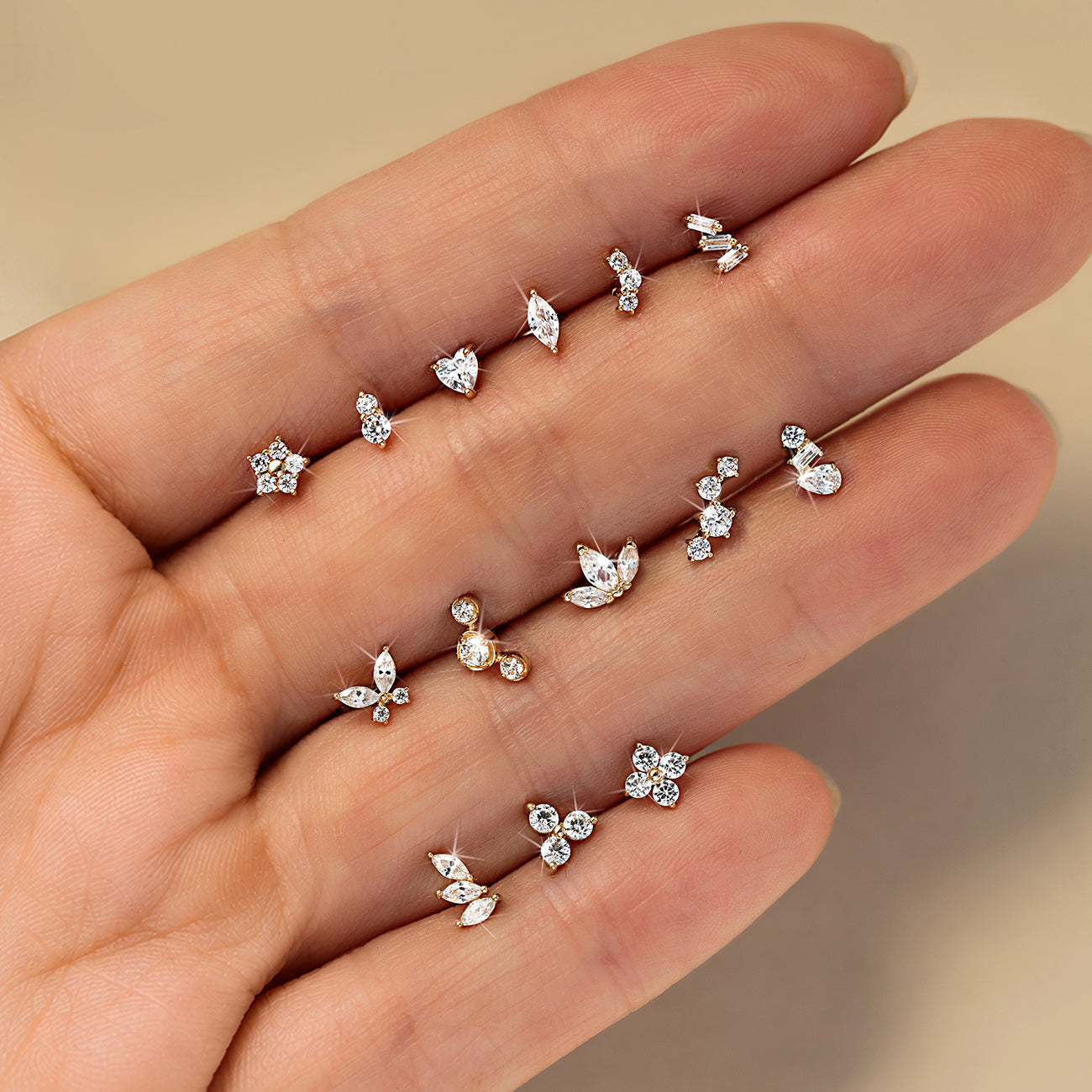 Fourteen gold flat back crystal studs in star, marquise, butterfly, cluster and baguette shapes resting in between fingers