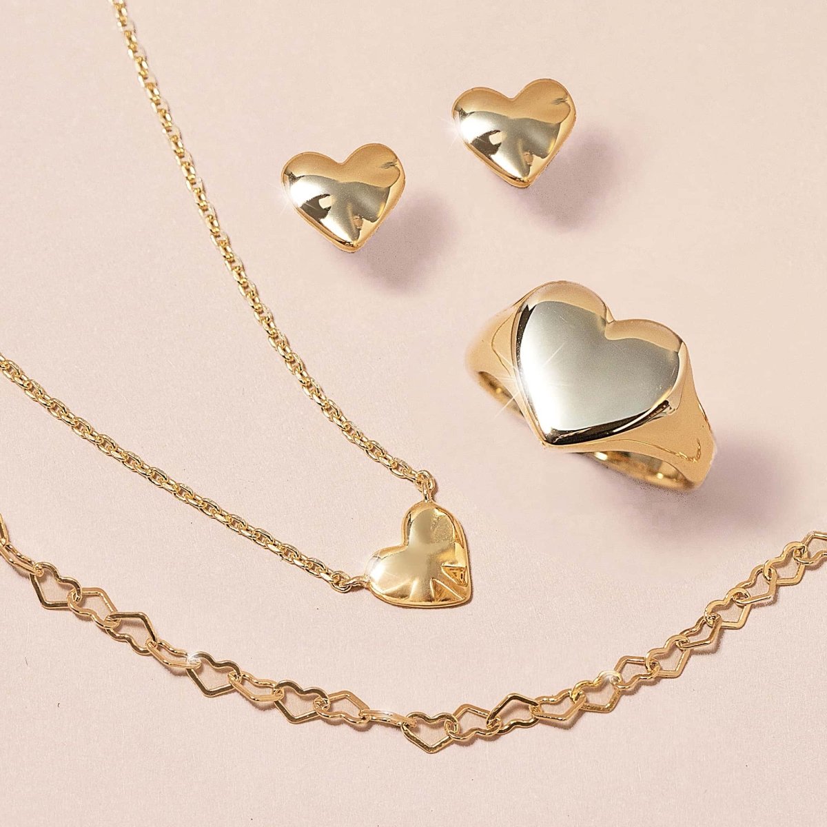 Gold-filled open heart-link chain bracelet shown with gold heart studs, ring and necklace on a cream background