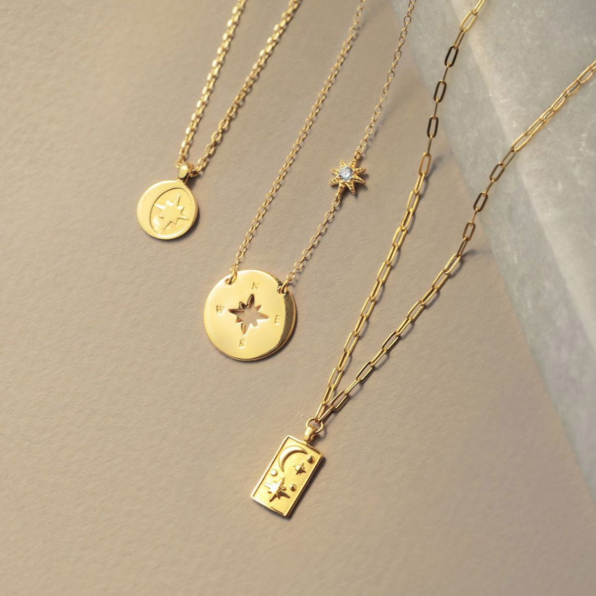 Three gold celestial pendant necklaces with compass-star discs and a moon-and-star tag, on a cream background