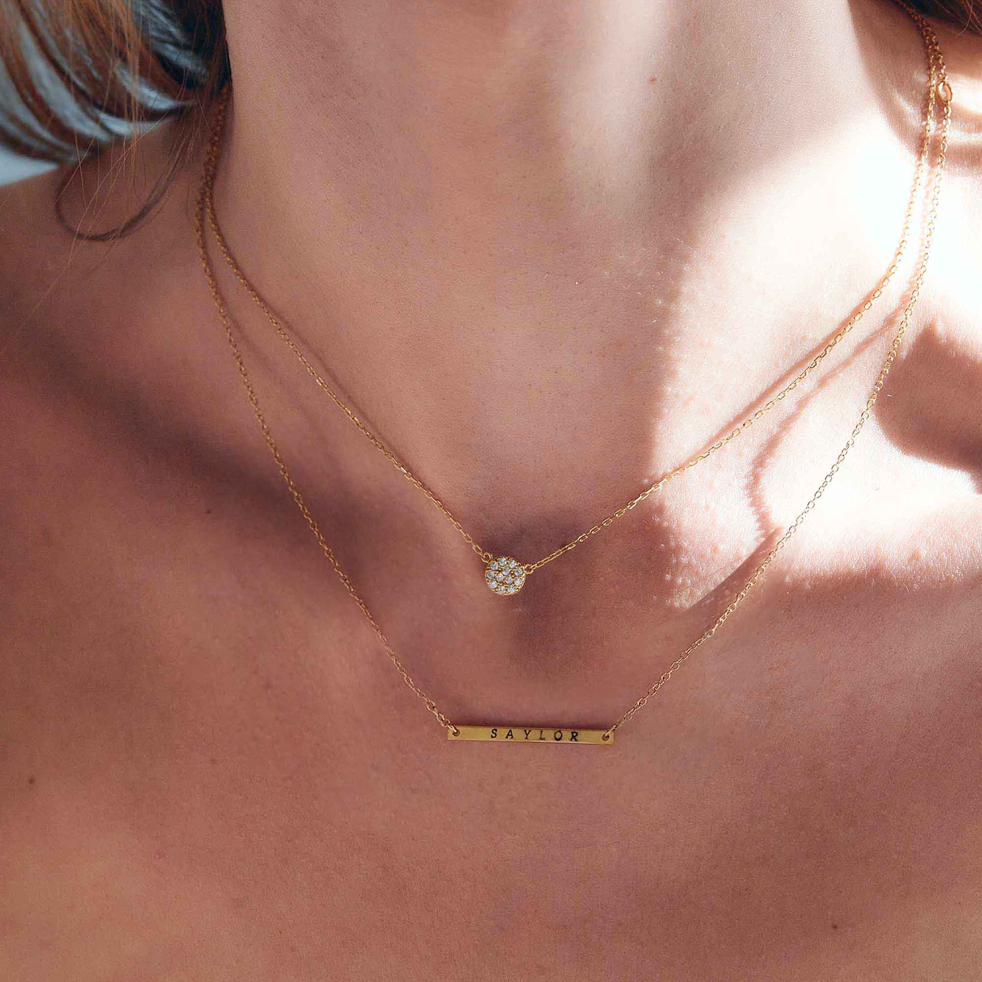 Gold-filled thin bar necklace engraved with a name, layered below a stone pendant on a model's neckline