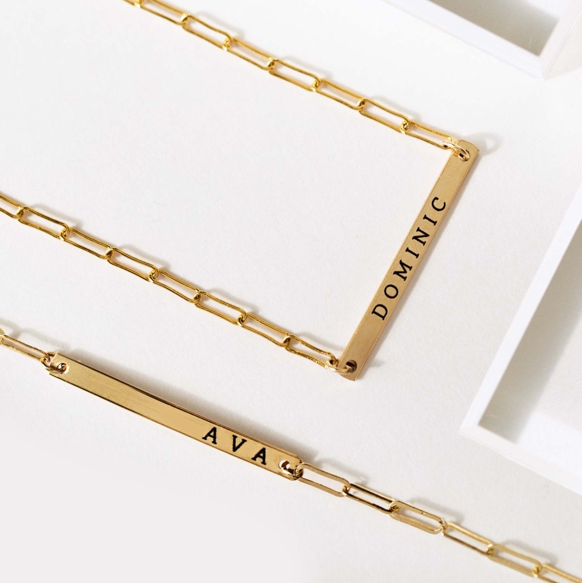 Gold-filled thin bar necklaces engraved with names, laid on a white surface, close-up detail