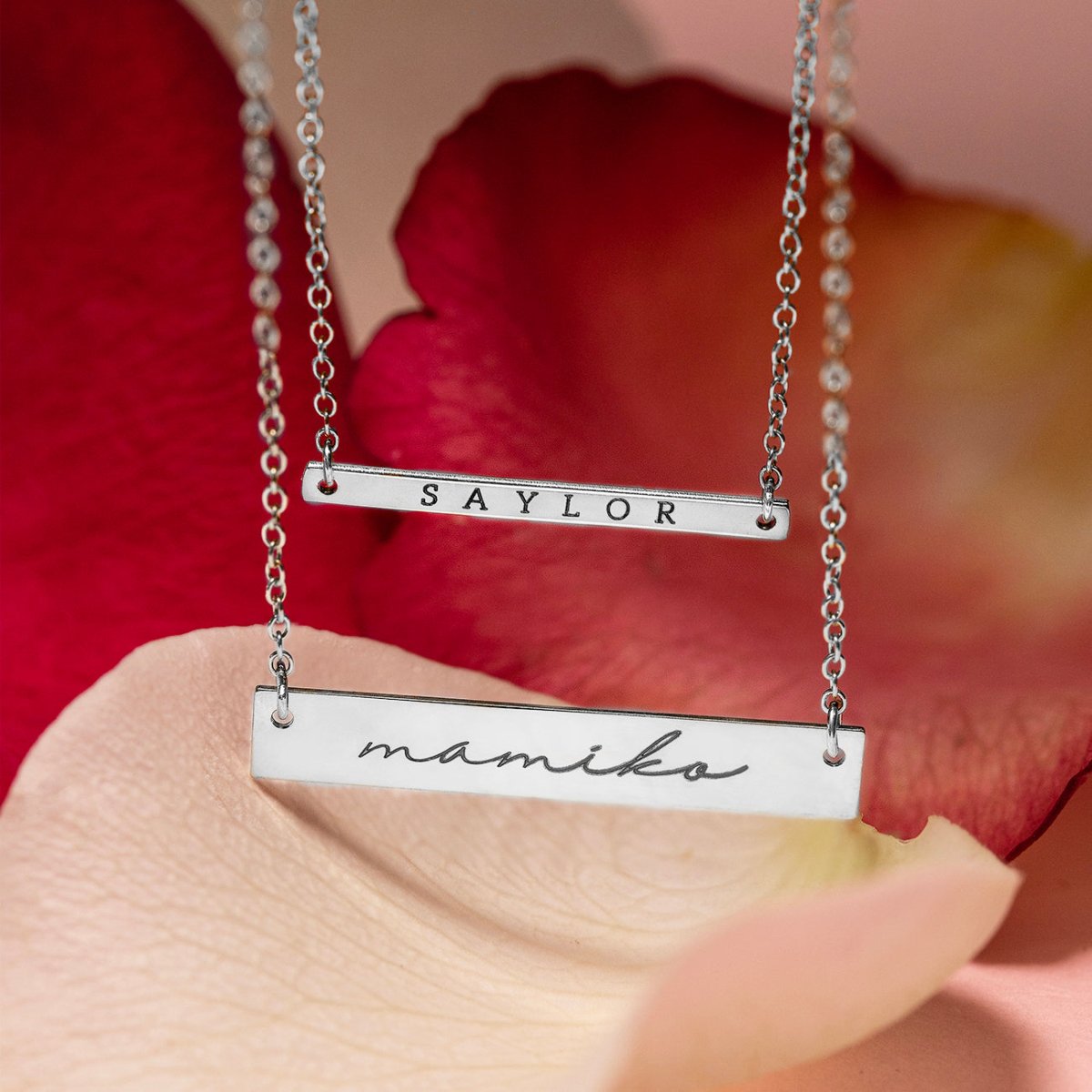 Two sterling silver thin bar necklaces engraved with names, resting on pink and red rose petals