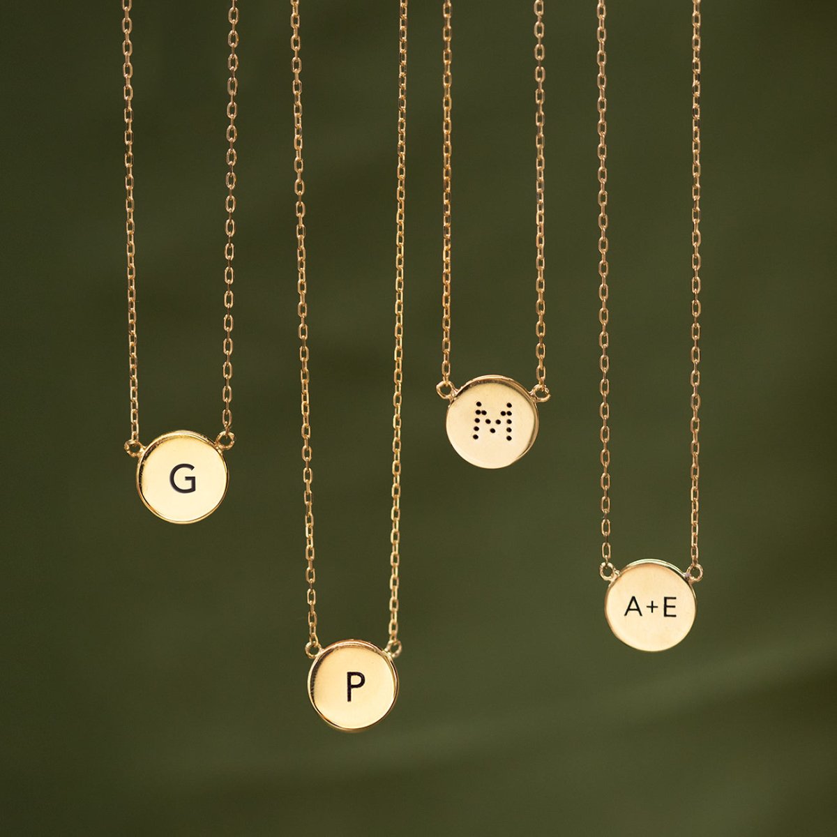Four gold vermeil disc necklaces each engraved with an initial, shown against a green background