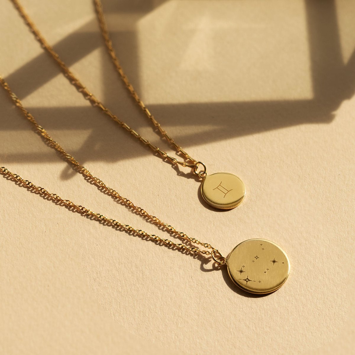 Two gold vermeil zodiac disc necklaces with engraved star symbols, laid on a cream background