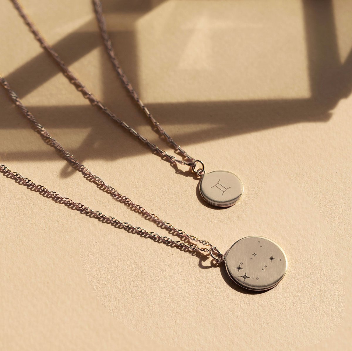 Two sterling silver disc necklaces, one with a zodiac symbol and one constellation, on a tan surface