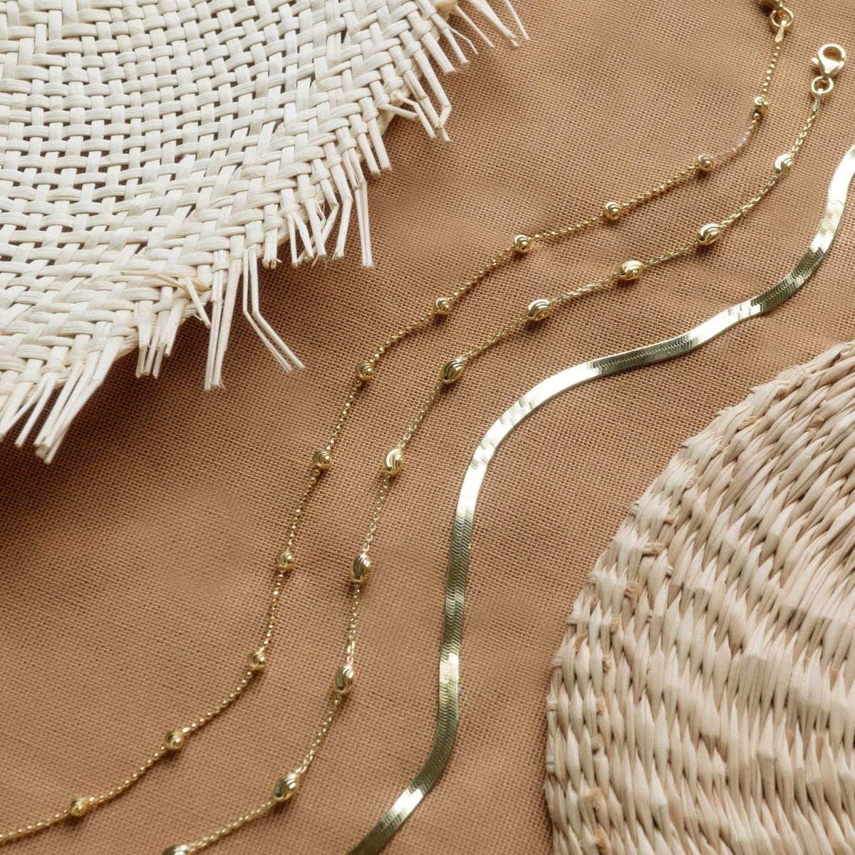 Gold vermeil faceted beaded anklet draped on tan cloth beside woven rattan props