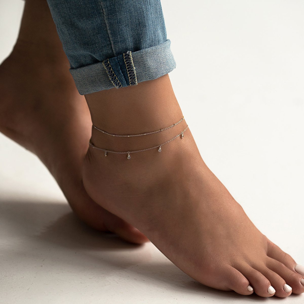 Sterling silver tiny clear-stone dangle anklet layered on a model's ankle with cuffed jeans