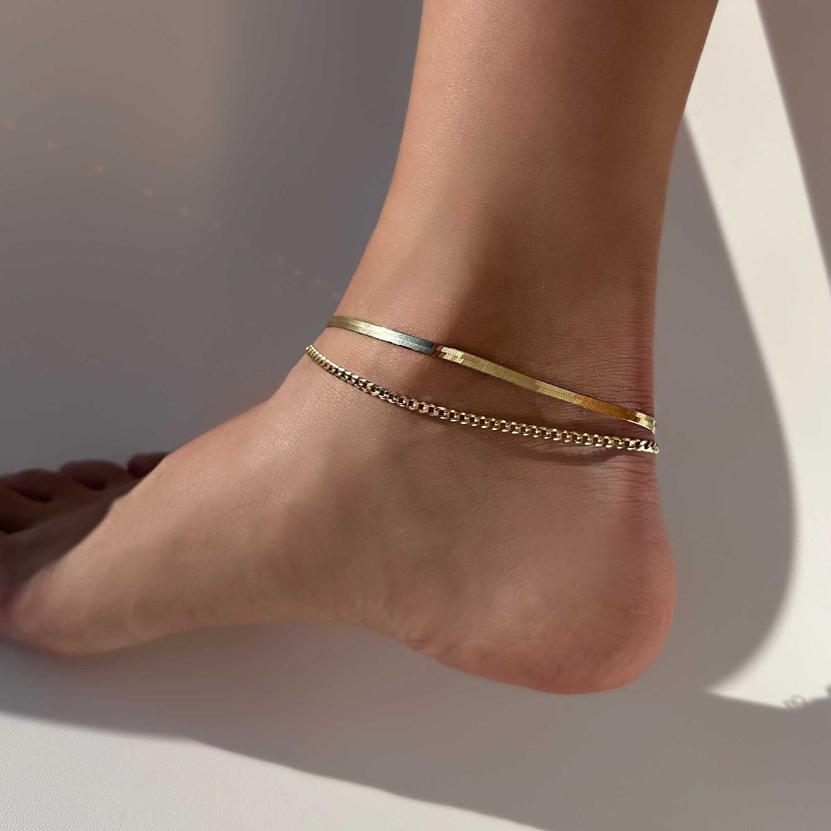14K gold flat Cuban curb chain anklet layered with a flat snake chain, shown on a model's ankle