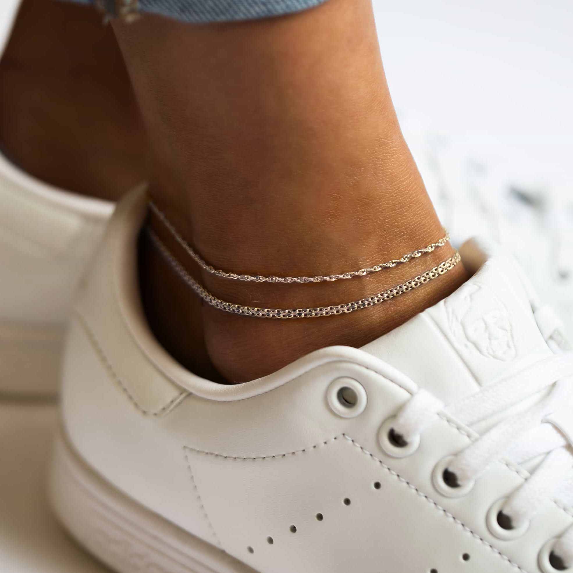 Two silver fine chain anklets layered on a model's ankle above a white sneaker