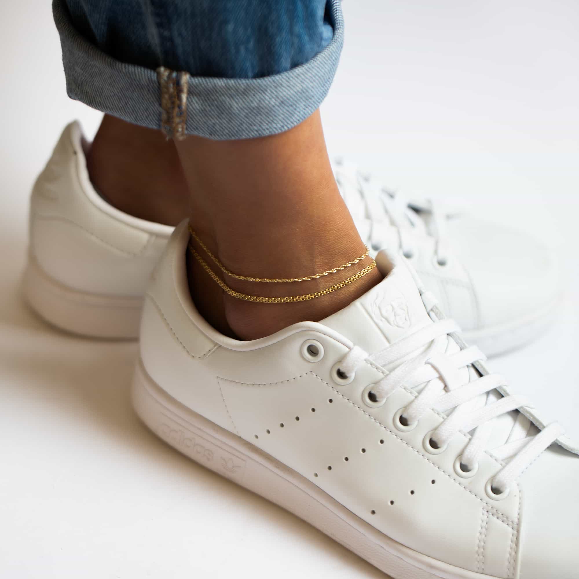 Two gold fine chain anklets on a model's ankle with white sneakers and cuffed jeans
