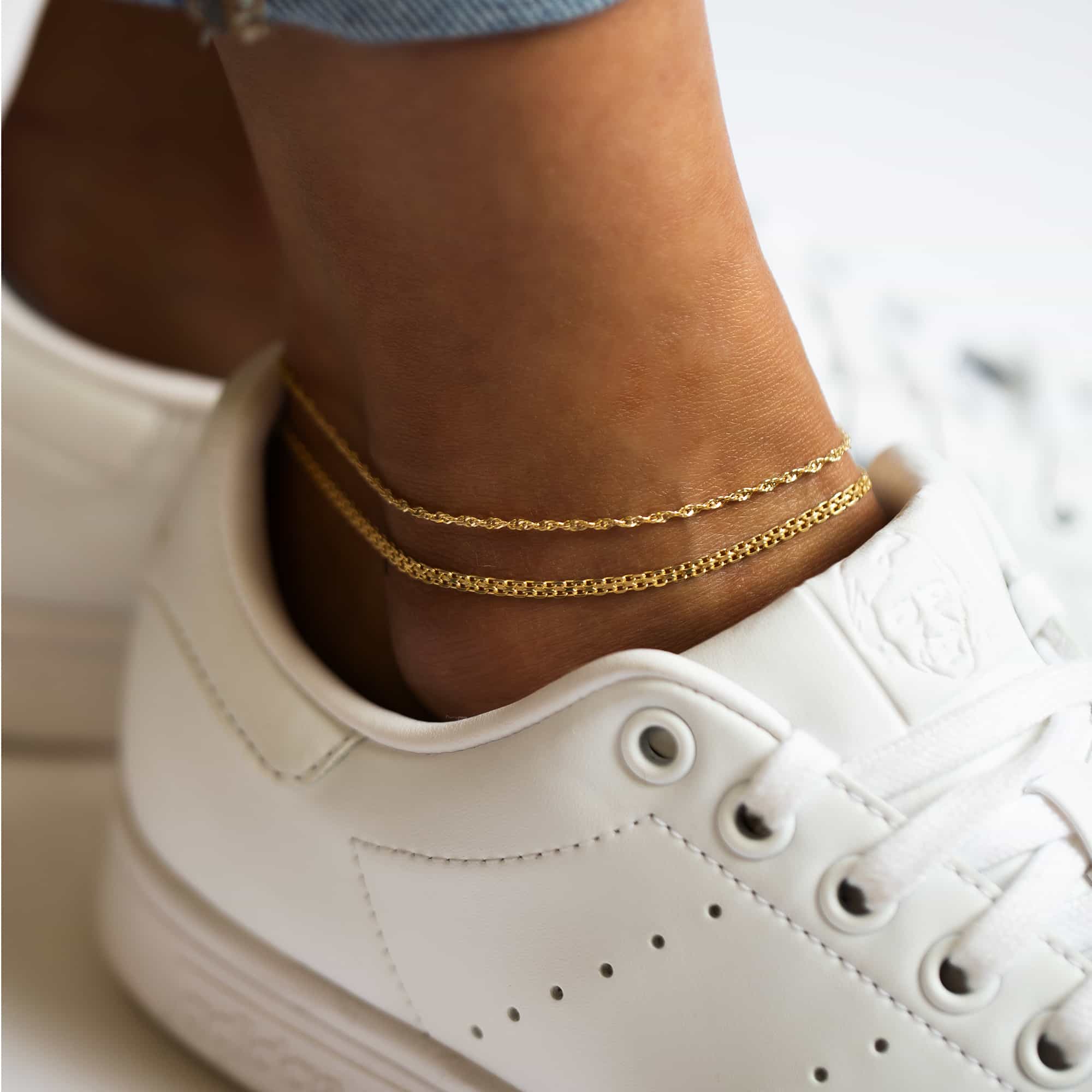 Two gold fine chain anklets layered on a model's ankle above a white sneaker
