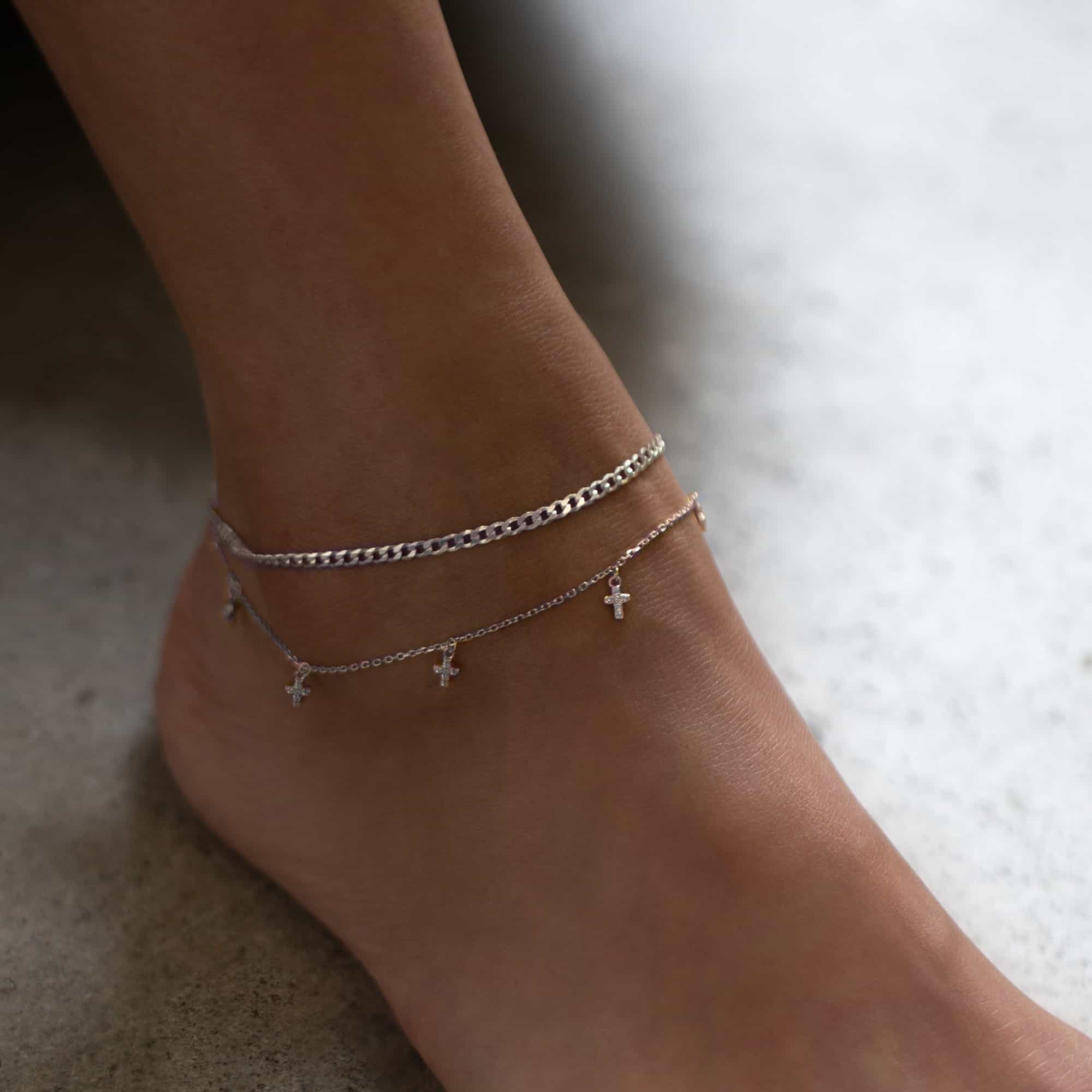 Silver curb chain anklet layered with a cross-charm anklet, shown close-up on a model's ankle