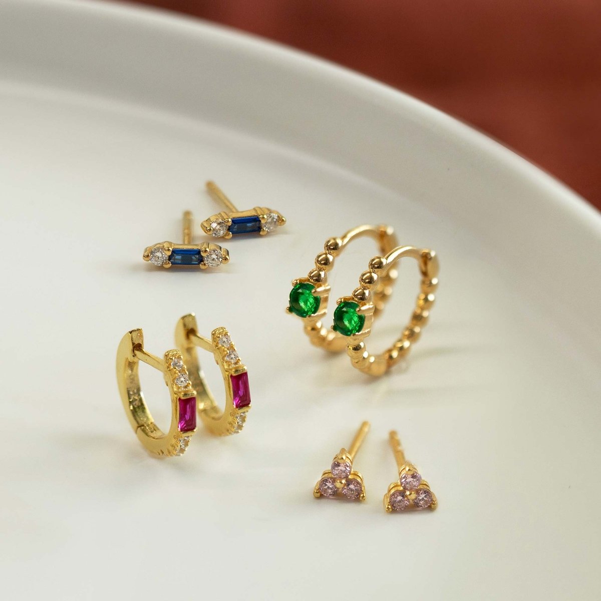 Gold vermeil baguette studs with blue stones, shown with other earrings on a white dish