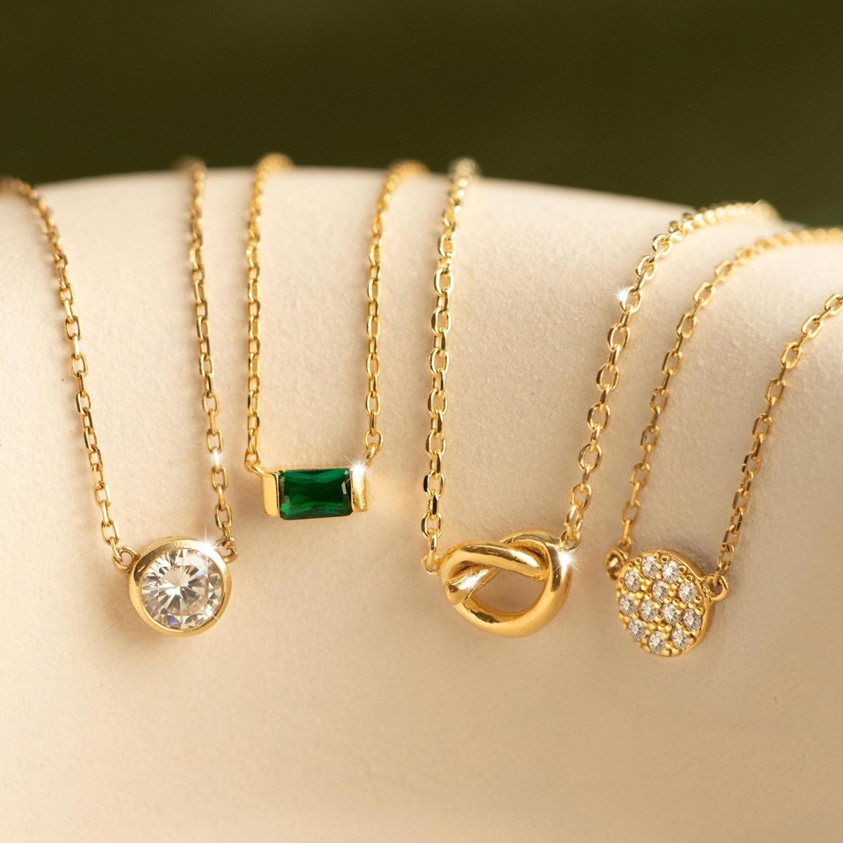 Gold vermeil emerald baguette necklace shown beside other gold pendants on a beige cushion, close-up detail