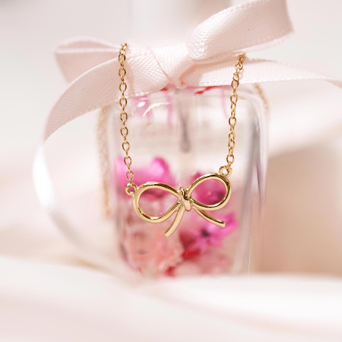 Gold vermeil bow necklace draped over a glass bottle with pink ribbon and dried flowers