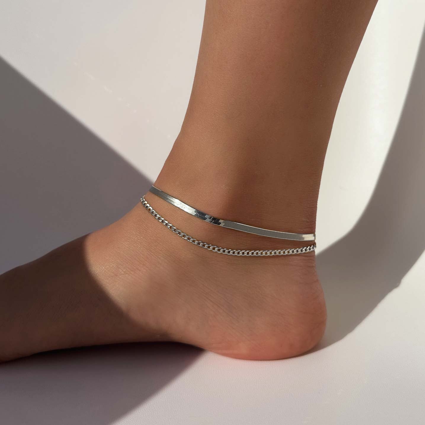Sterling silver flat Cuban curb chain anklet layered with a flat snake chain, shown on a model's ankle