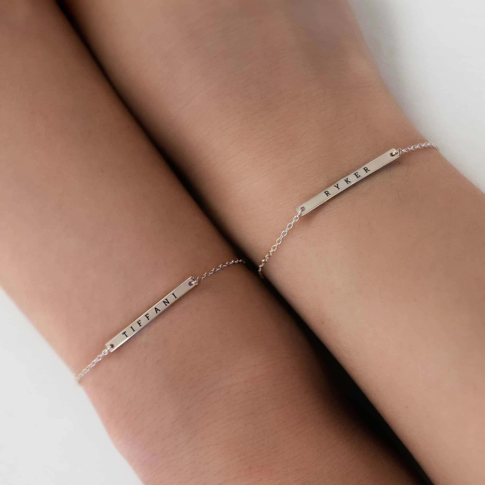 Two sterling silver thin bar bracelets engraved with names, shown on crossed wrists