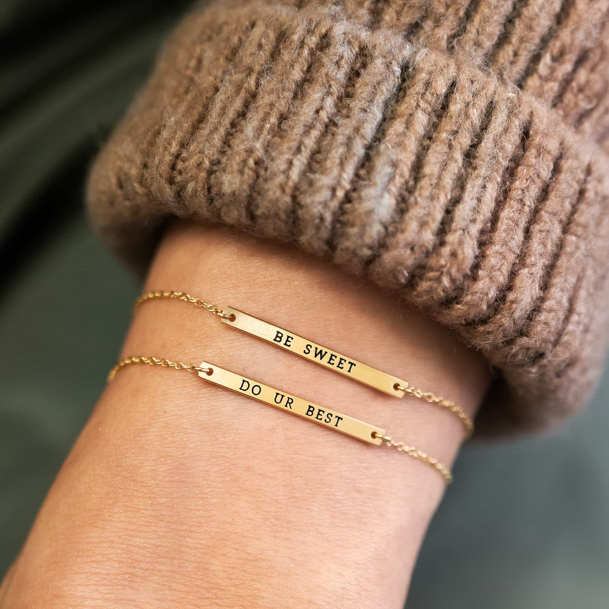 Two gold-filled thin bar bracelets engraved with phrases, stacked on a model's wrist by a sweater