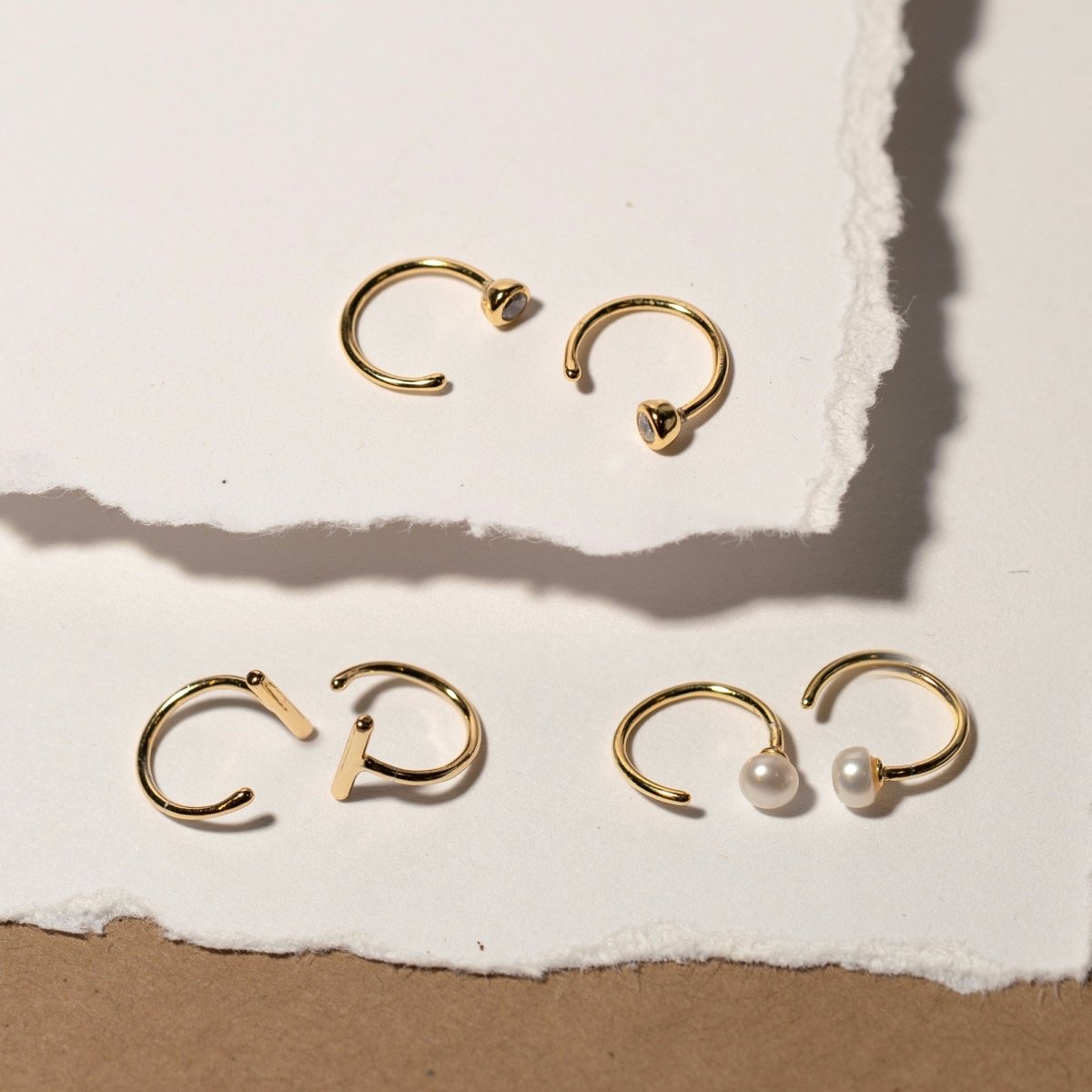 Gold vermeil bar huggie earrings with clear stone and pearl variants, flat-laid on torn paper