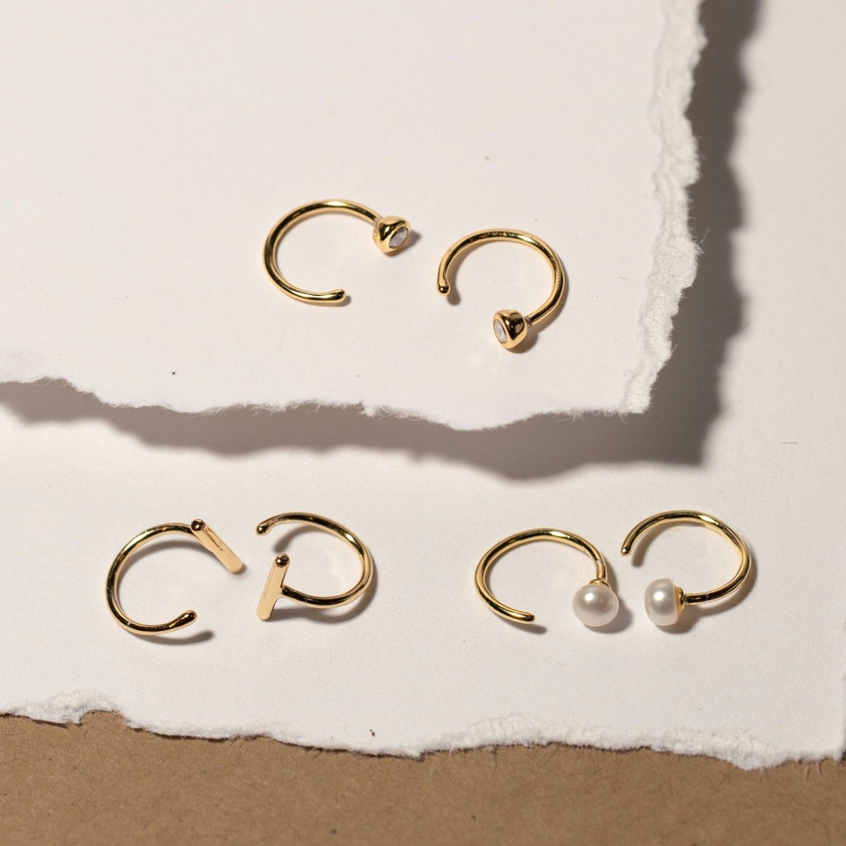 Gold open hug hoops with pearls beside a clear-stone pair and a bar pair, on torn paper