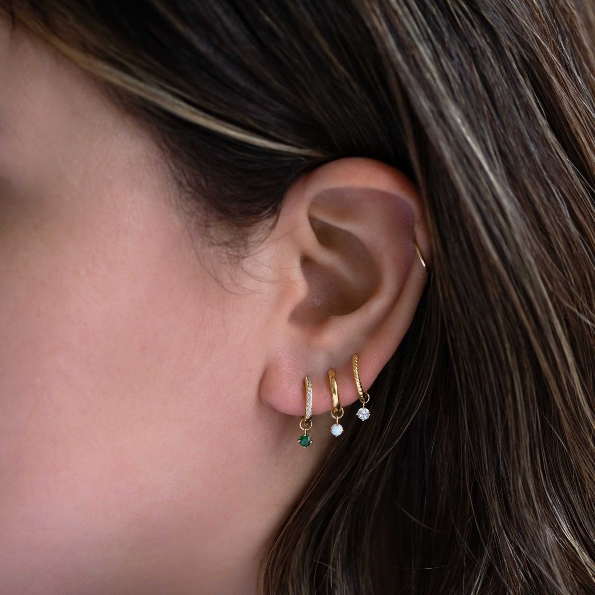 14K gold round emerald charm hanging from a huggie hoop, shown on a model's lobe