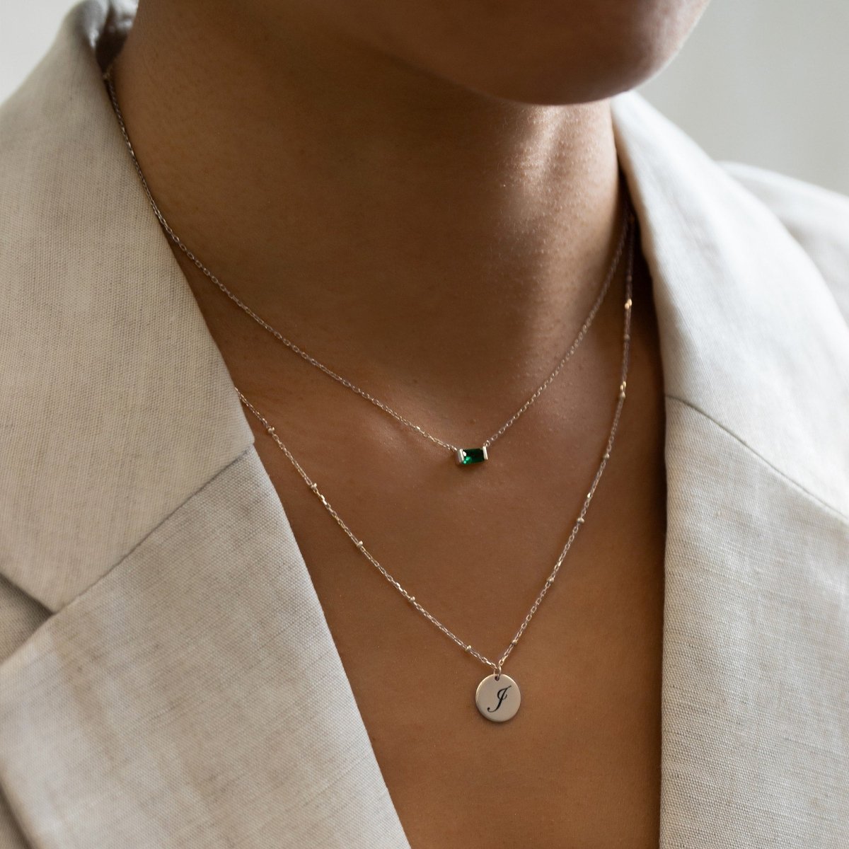 Sterling silver emerald baguette necklace layered with an initial disc pendant, shown on a model's neckline